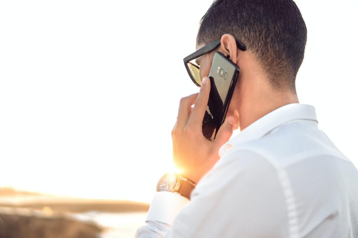 Hombre con camisa blanca y gafas de sol sosteniendo un teléfono en su oreja al aire libre, mientras el sol se pone detrás de él.