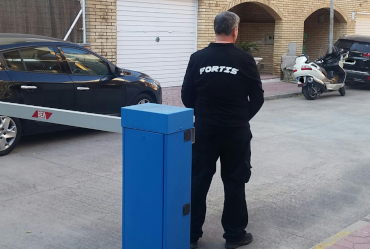 Un guardia de seguridad con uniforme negro está junto a una barrera azul. Cerca hay un coche negro en un remolque.