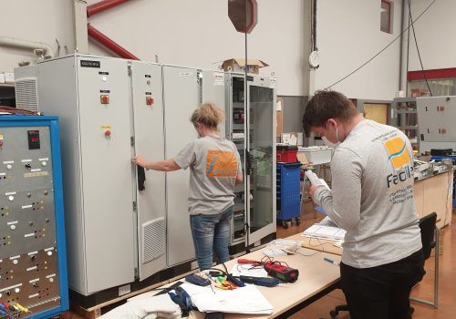 Deux personnes portant des chemises grises avec des logos, travaillant sur des panneaux électriques dans un atelier.