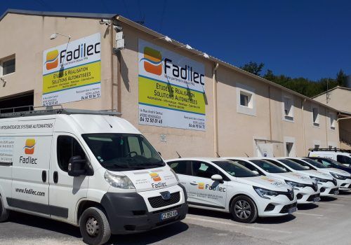 Des fourgonnettes et des voitures blanches Fadilec garées devant un bâtiment portant des panneaux Fadilec.
