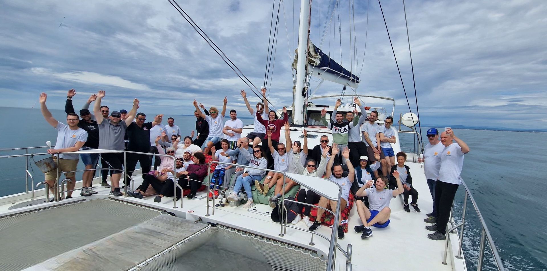 Groupe de personnes sur le pont d'un voilier, levant les bras en signe de célébration par une journée nuageuse.