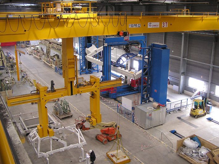 Vue aérienne d'une usine avec une grue jaune, des machines bleues et des ouvriers.