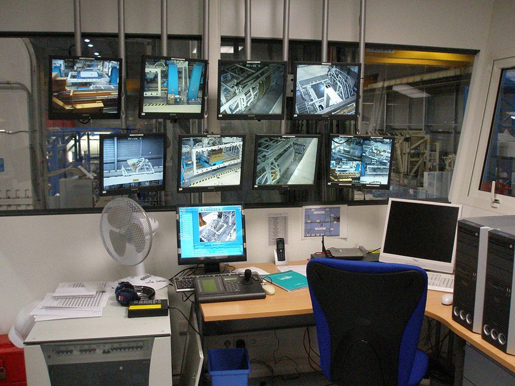 Salle de contrôle avec plusieurs écrans affichant des vues de l'usine ; bureau avec ordinateurs et ventilateur.