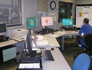 Salle de contrôle : homme à son bureau, écrans avec données, imprimante, horloge, espace de travail organisé.