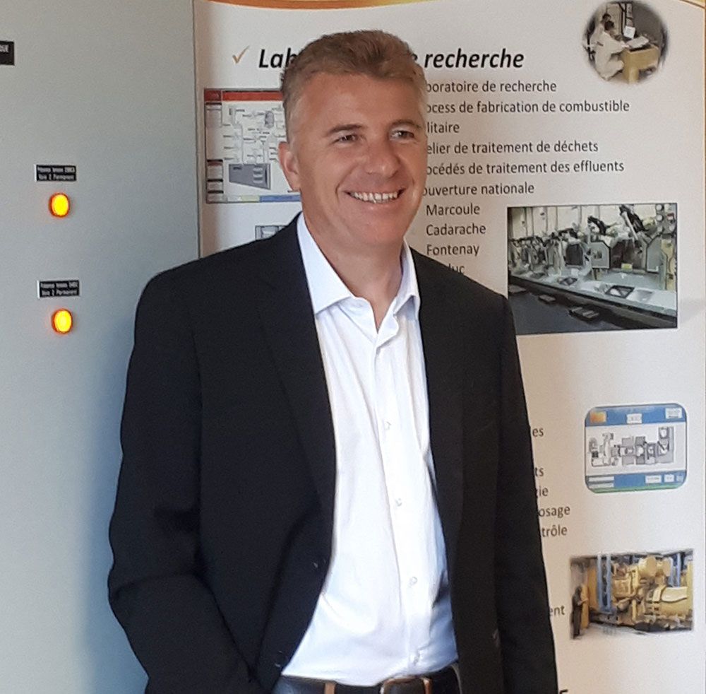 Un homme en blazer sourit, debout près d'un panneau et d'une affiche de recherche, probablement dans un laboratoire.