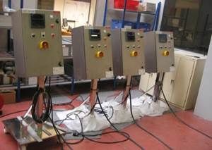 Quatre panneaux de contrôle gris avec boutons jaunes, posés sur pieds, dans un décor d'atelier.