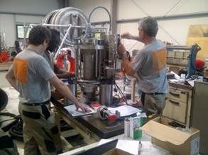 Trois hommes travaillent sur des machines dans un atelier, l'un d'eux utilise un outil, examine des papiers et assemble des pièces.
