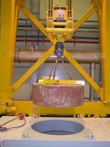 Une grue jaune soulevant un grand objet cylindrique au-dessus d'une ouverture ronde dans une plate-forme grise.