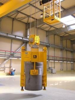 Pont roulant jaune soulevant un grand cylindre gris à l'intérieur d'un bâtiment industriel.