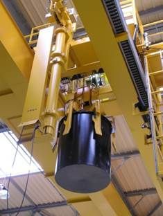 Pont roulant jaune soulevant un grand conteneur en métal noir dans un environnement industriel.