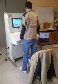 Homme utilisant une machine, vêtu d'une veste beige et noire et d'un jean. Veste supplémentaire sur une chaise ; ordinateur portable sur une table à côté.