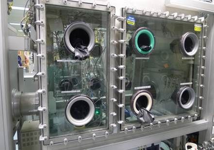 Deux boîtes à gants en verre connectées avec des ports de gants noirs, bleus et verts pour la manipulation de matériaux sensibles dans un environnement contrôlé.