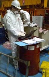 Une personne en combinaison de protection inspecte des documents au sommet d'un baril rouge dans un environnement industriel.