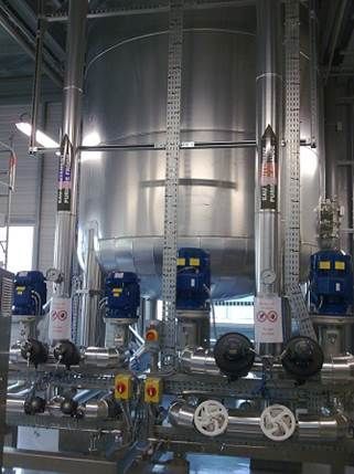 Grand réservoir industriel en acier inoxydable avec tuyaux, pompes, jauges et échelles à l'intérieur d'une usine.