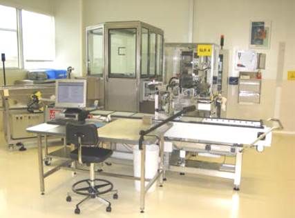 Un poste de travail industriel avec une machine, un ordinateur et une chaise. Environnement lumineux avec sol blanc.