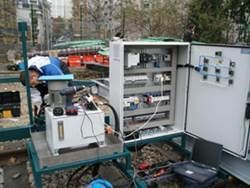 Un homme travaille sur une machine près d'un panneau de commande à l'extérieur, peut-être sur une voie ferrée.