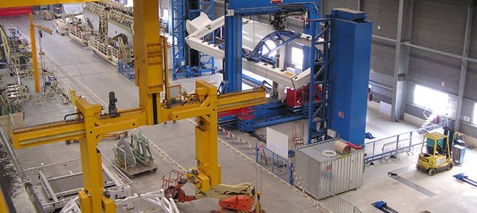 Une usine de fabrication industrielle avec des grues jaunes, des machines bleues et un petit chariot élévateur jaune.