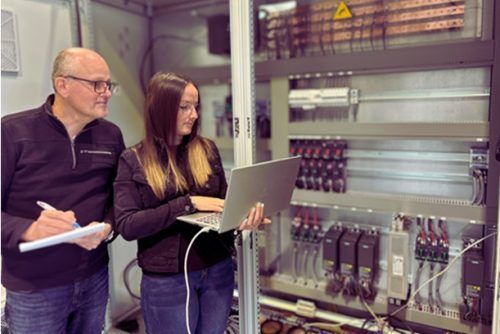 Deux personnes, l’une avec un ordinateur portable et l’autre avec un bloc-notes, examinent des équipements électriques dans un environnement industriel.