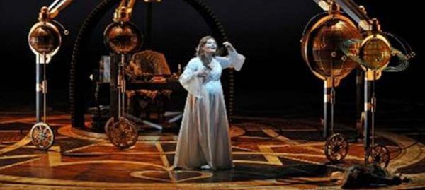 Femme en robe blanche chantant sur une scène avec des machines de style steampunk.