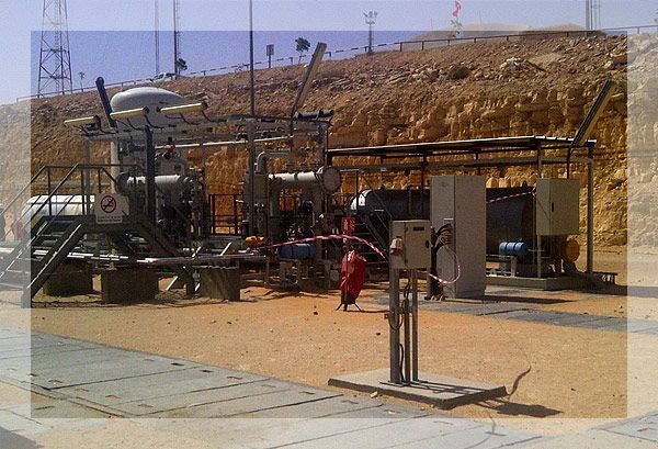 Équipement industriel sur un terrain rocailleux et désertique. Structures métalliques, tuyaux et boîtier électrique sont visibles.