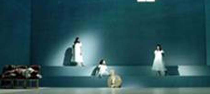 Trois femmes en robes blanches se tiennent sur une scène bleu sarcelle. Un projecteur les braque sur elles.