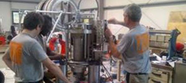 Trois personnes travaillent sur des machines dans un atelier. Elles portent des t-shirts gris à imprimé orange.