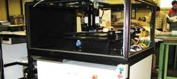 Une grande machine avec une enceinte en verre, à l'intérieur d'un atelier ; les gens sont à l'arrière-plan.