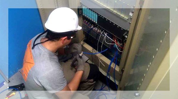 Un homme portant un casque et des gants travaille sur des câbles réseau dans un rack de serveur.