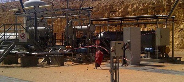 Équipement industriel dans une zone sablonneuse, probablement une installation pétrolière ou gazière. Une personne en rouge passe devant.