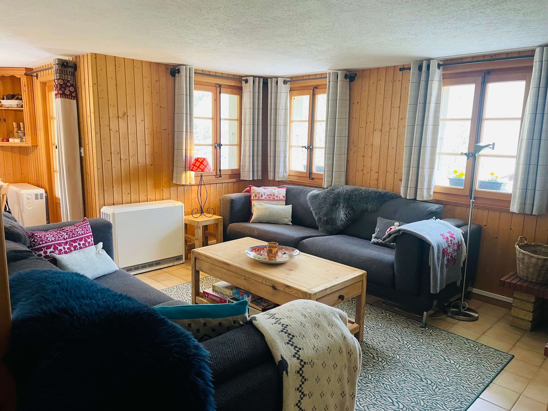 Salon cosy avec murs lambrissés, canapés gris et petites fenêtres à rideaux.