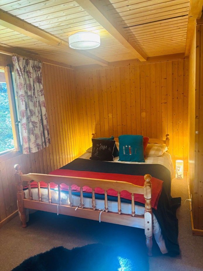 Chambre en bois avec lit, couverture rouge, oreillers et fenêtre avec rideaux à fleurs.