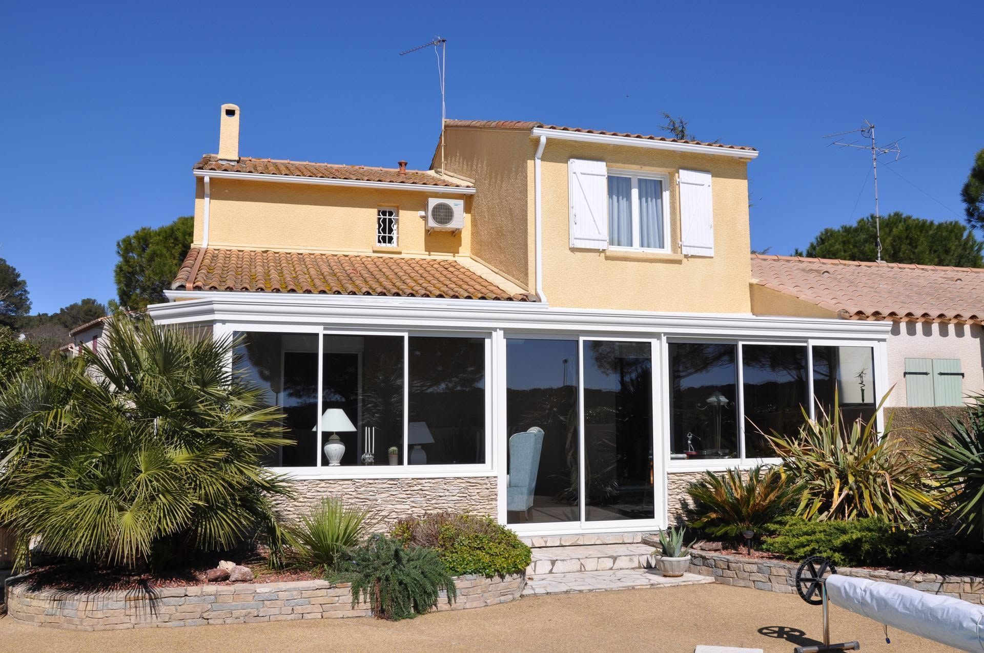 Maison en stuc baignée de soleil, avec toit de tuiles, grandes fenêtres et petit jardin sous un ciel bleu limpide.