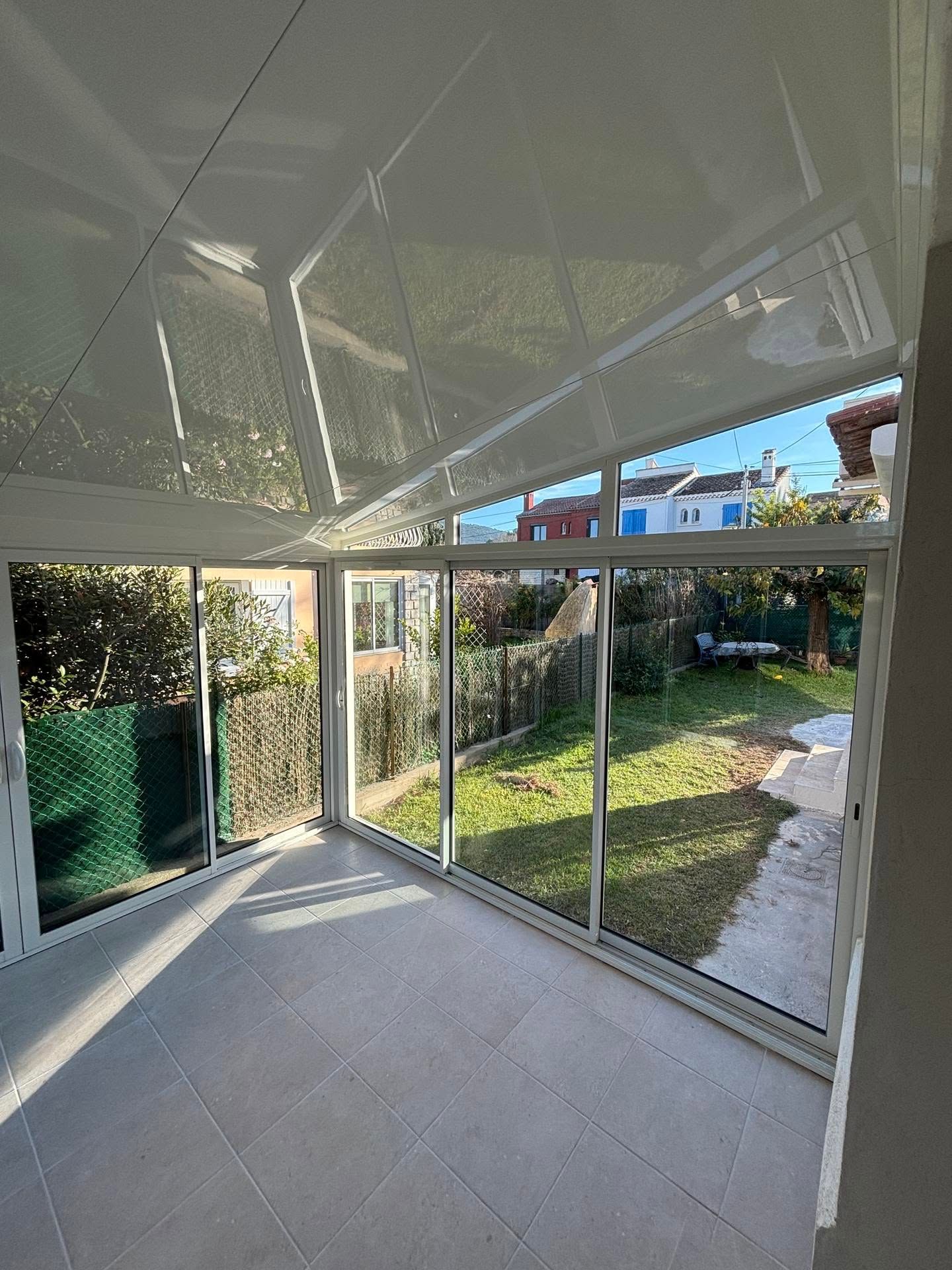 Véranda avec sol carrelé, parois vitrées et vue sur un jardin gazonné et les maisons voisines