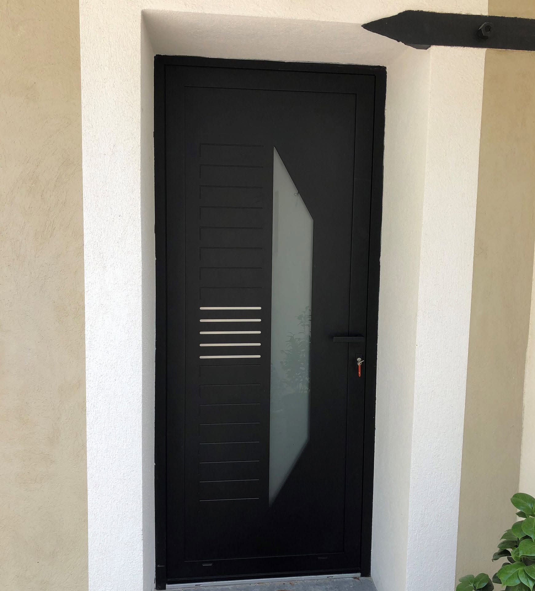 Porte d'entrée noire avec un panneau étroit en verre dépoli et des grilles d'aération horizontales dans un mur blanc.
