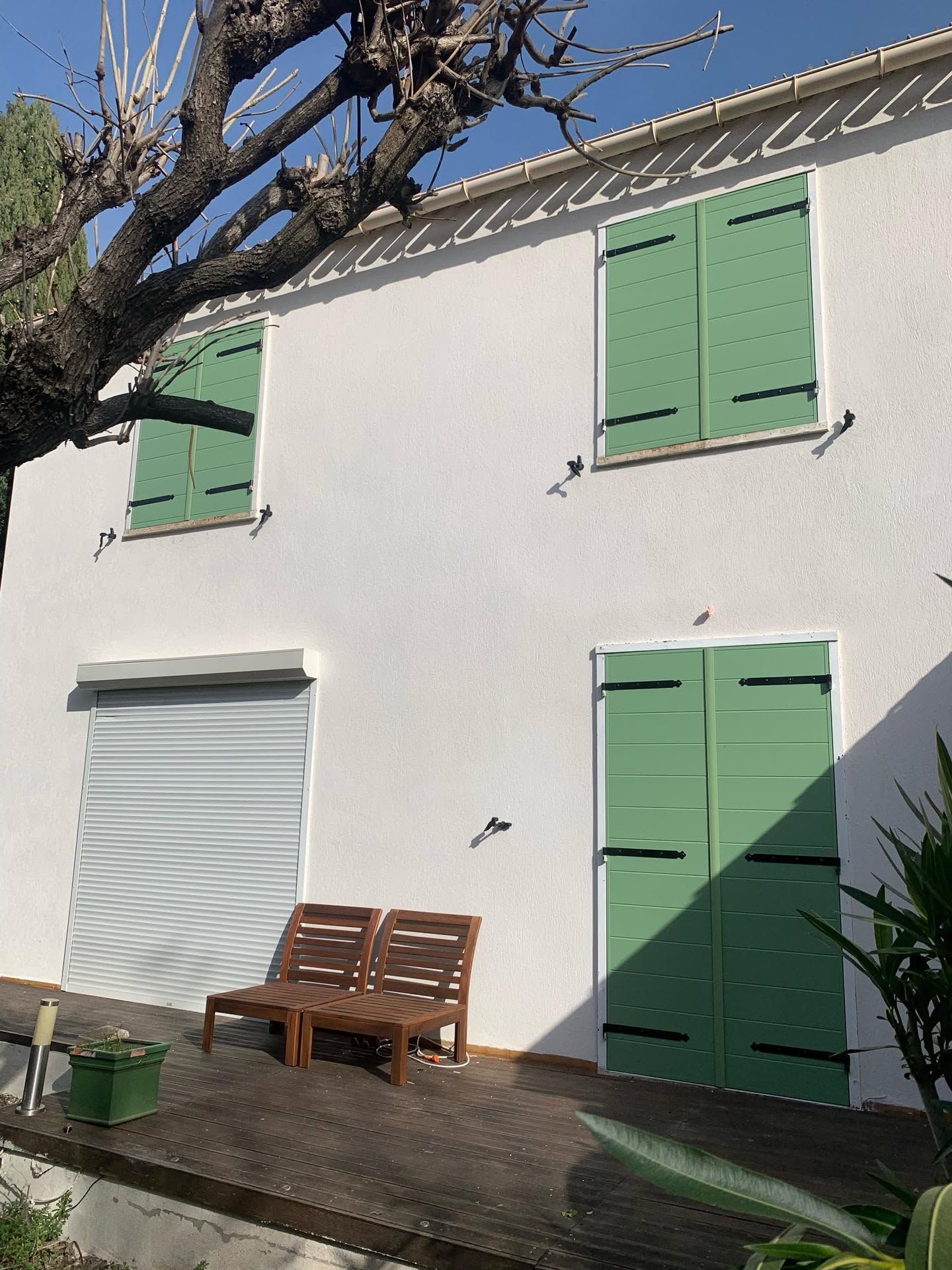 Extérieur d'une maison blanche avec des volets verts, une porte sectionnelle fermée et un banc en bois au soleil.