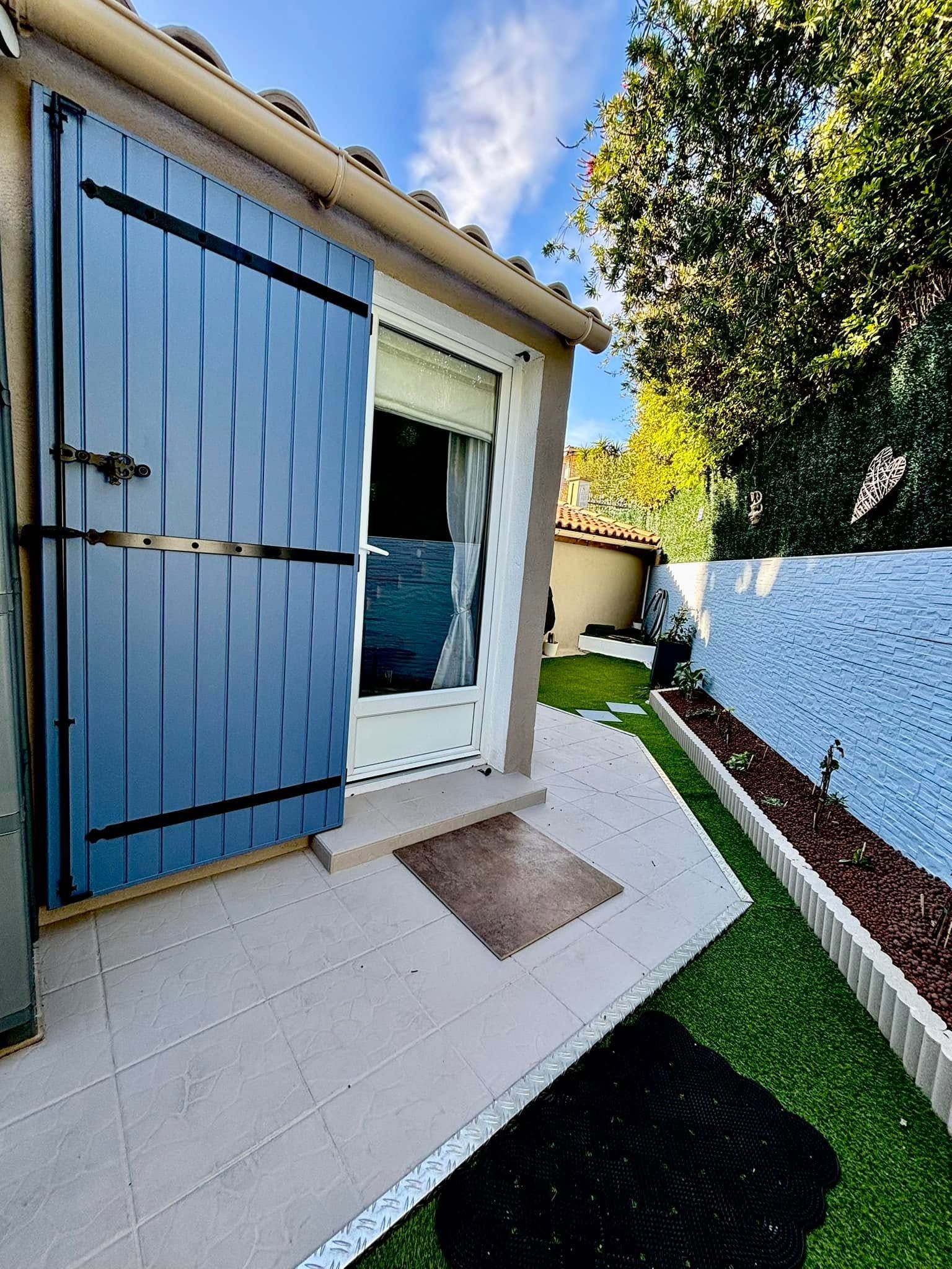 Porte d'entrée à volets bleus à côté d'un mur blanc et d'un porche carrelé, avec un petit jardin et un chemin.