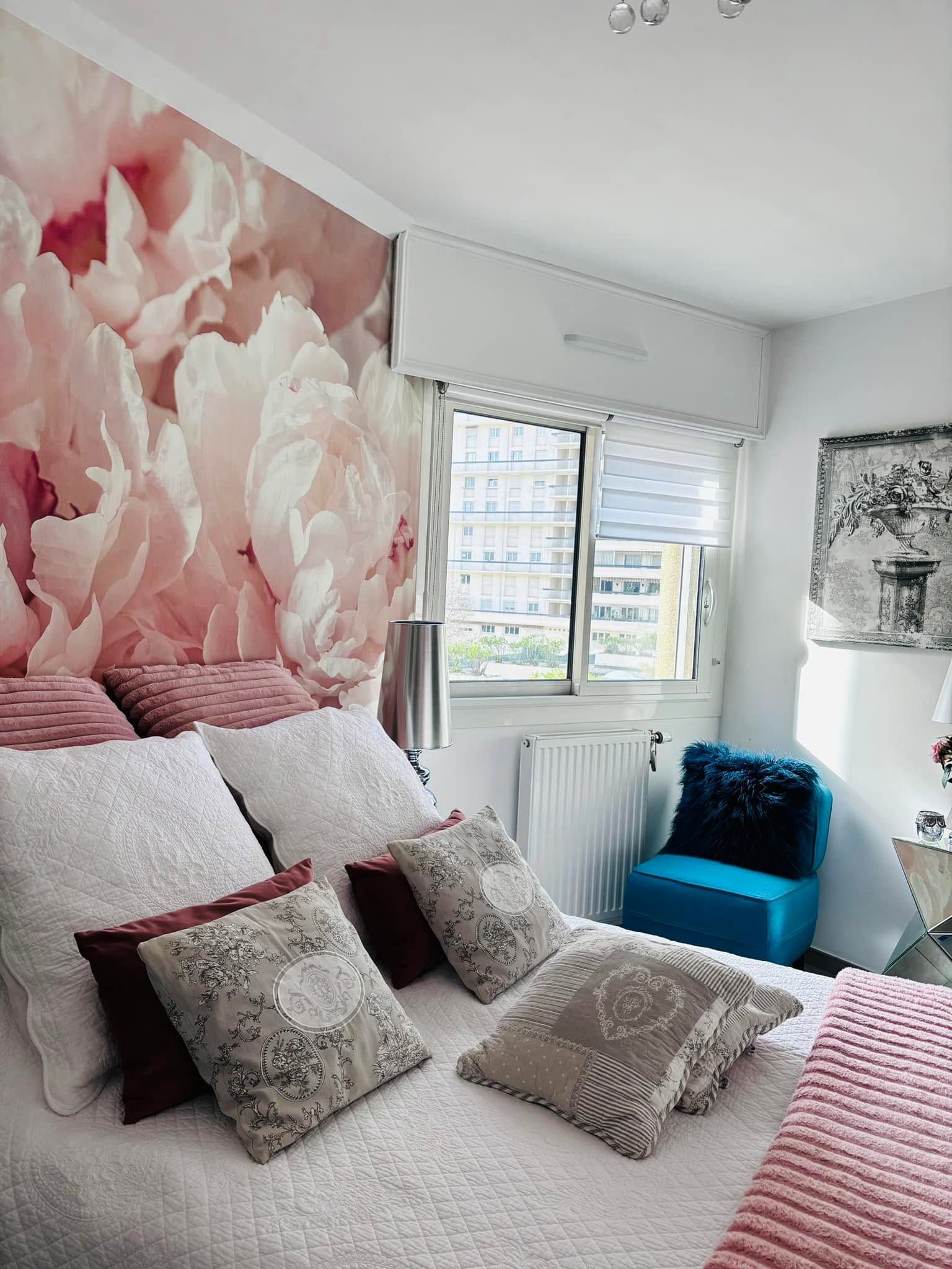 Chambre cosy avec une fresque murale florale rose, du linge de lit blanc et une chaise bleue près de la fenêtre.