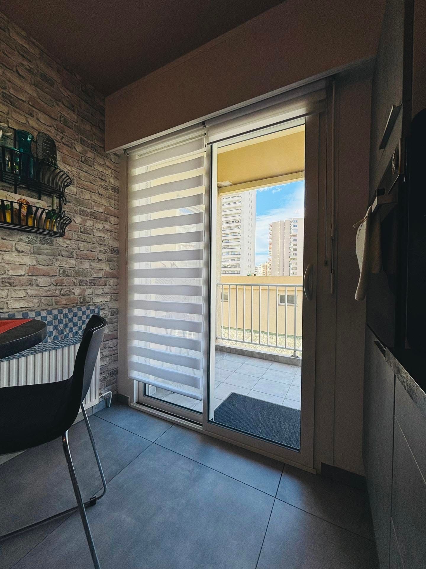 Vue depuis le balcon à travers une porte coulissante ouverte, avec des stores transparents et les immeubles d'appartements voisins visibles.