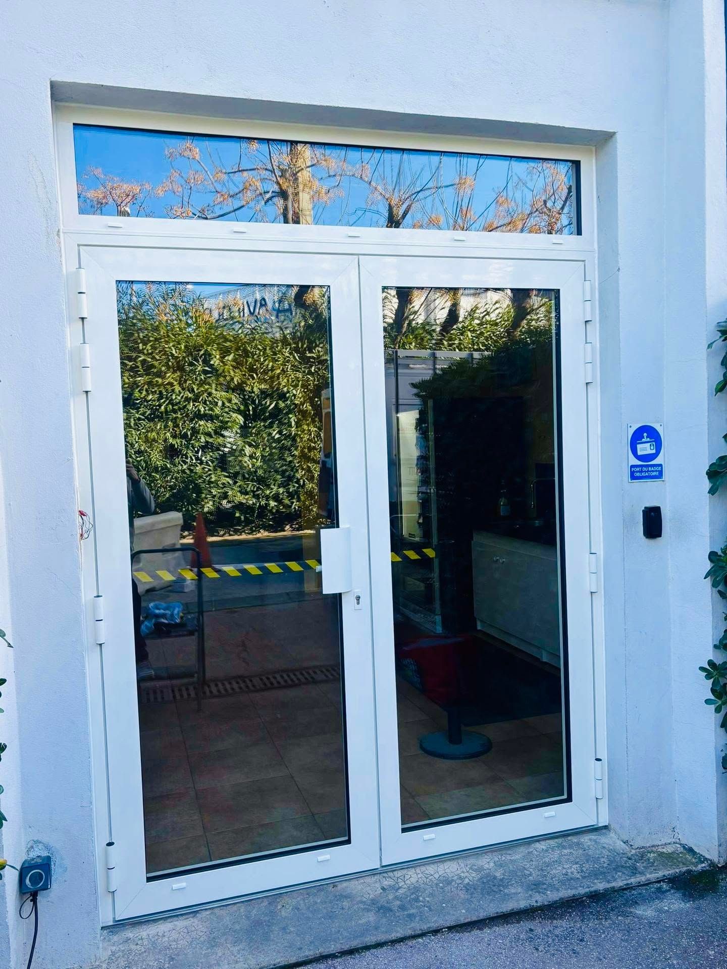 Double porte vitrée blanche à l'entrée d'un immeuble bleu, avec des arbres se reflétant dans le verre.