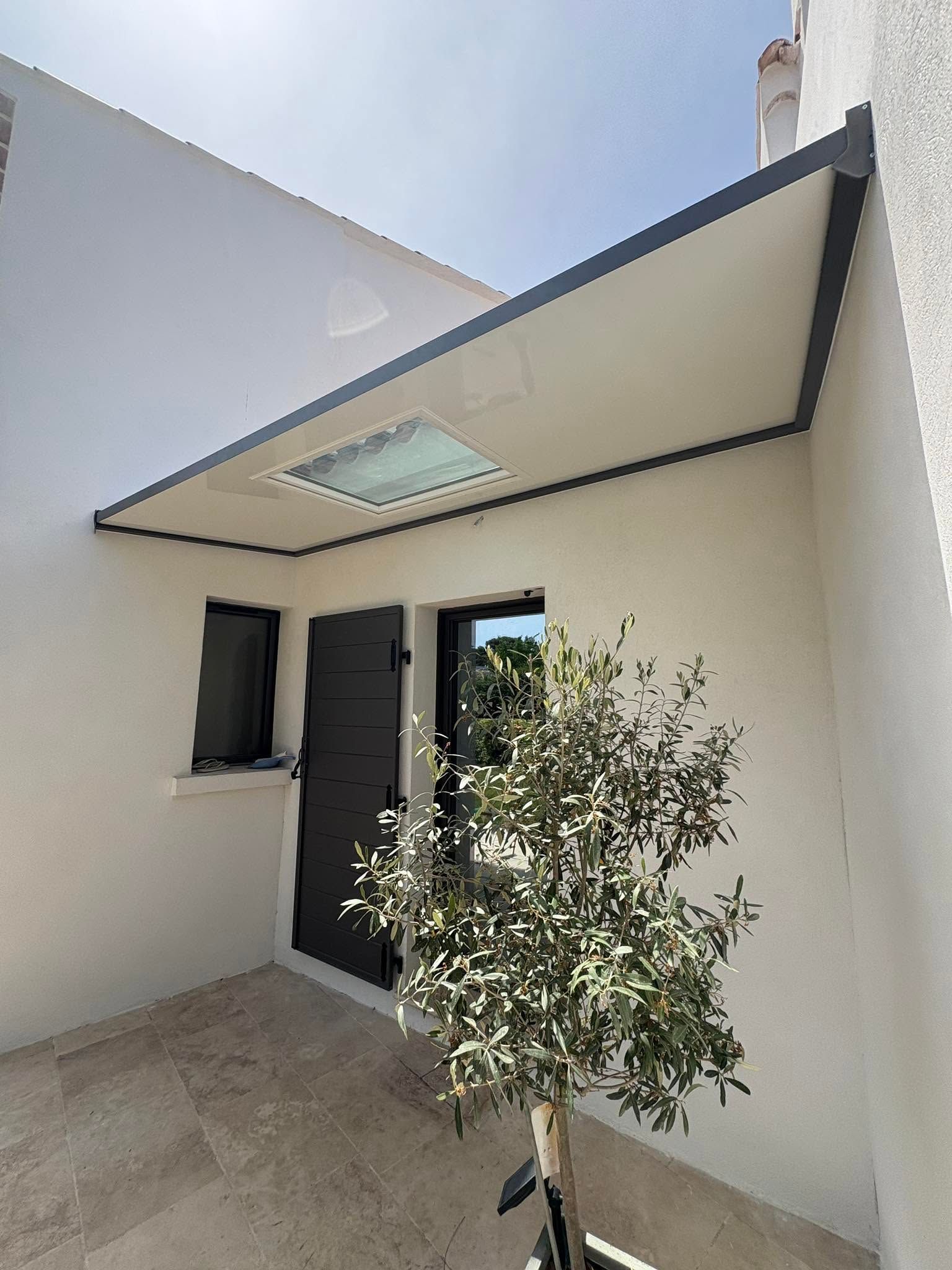 Entrée d'une maison blanche moderne avec porte noire, petite fenêtre et arbre en pot sous un auvent.