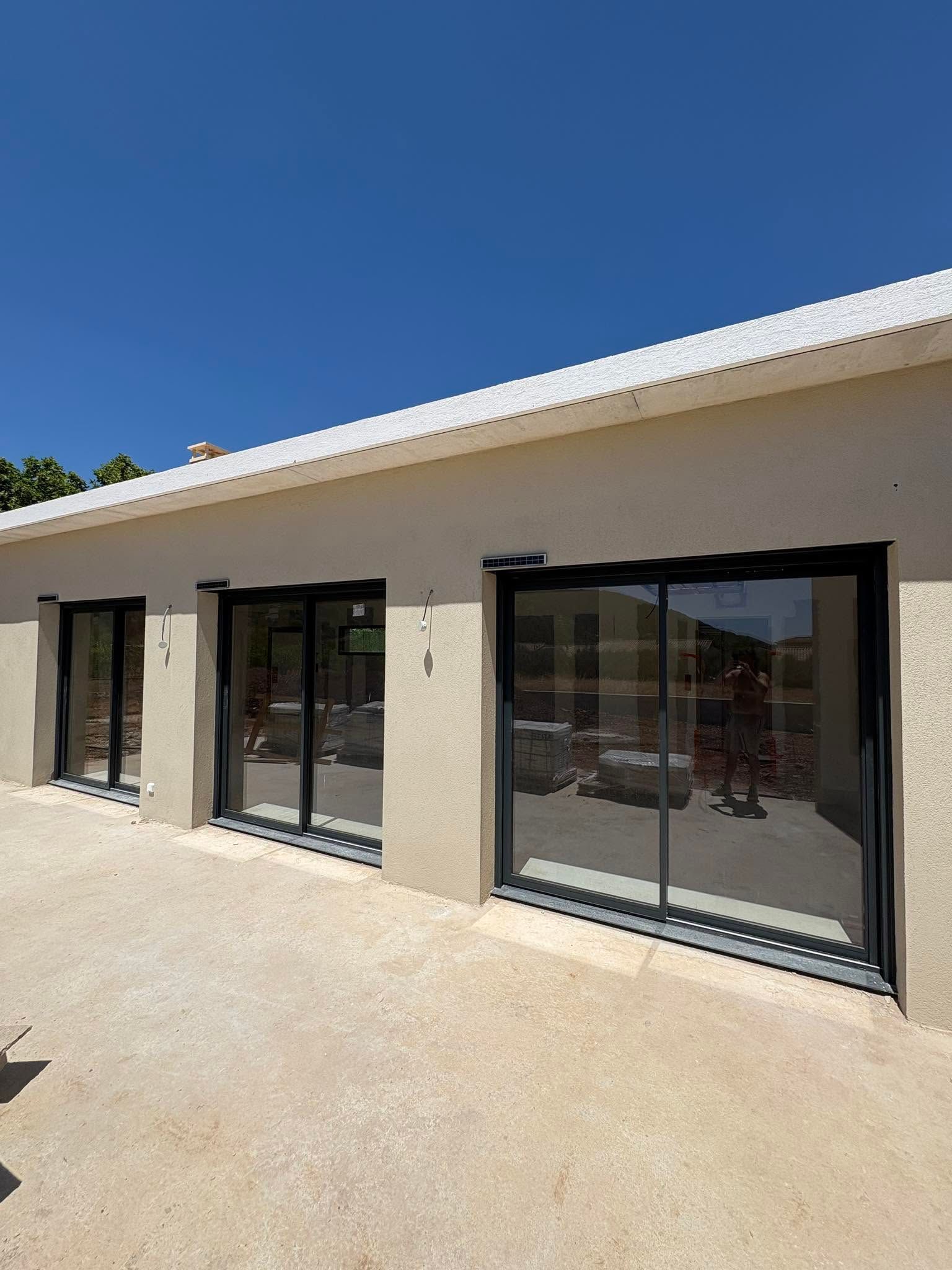 Façade beige moderne d'un bâtiment avec portes et fenêtres vitrées à cadre noir, sous un ciel bleu clair.