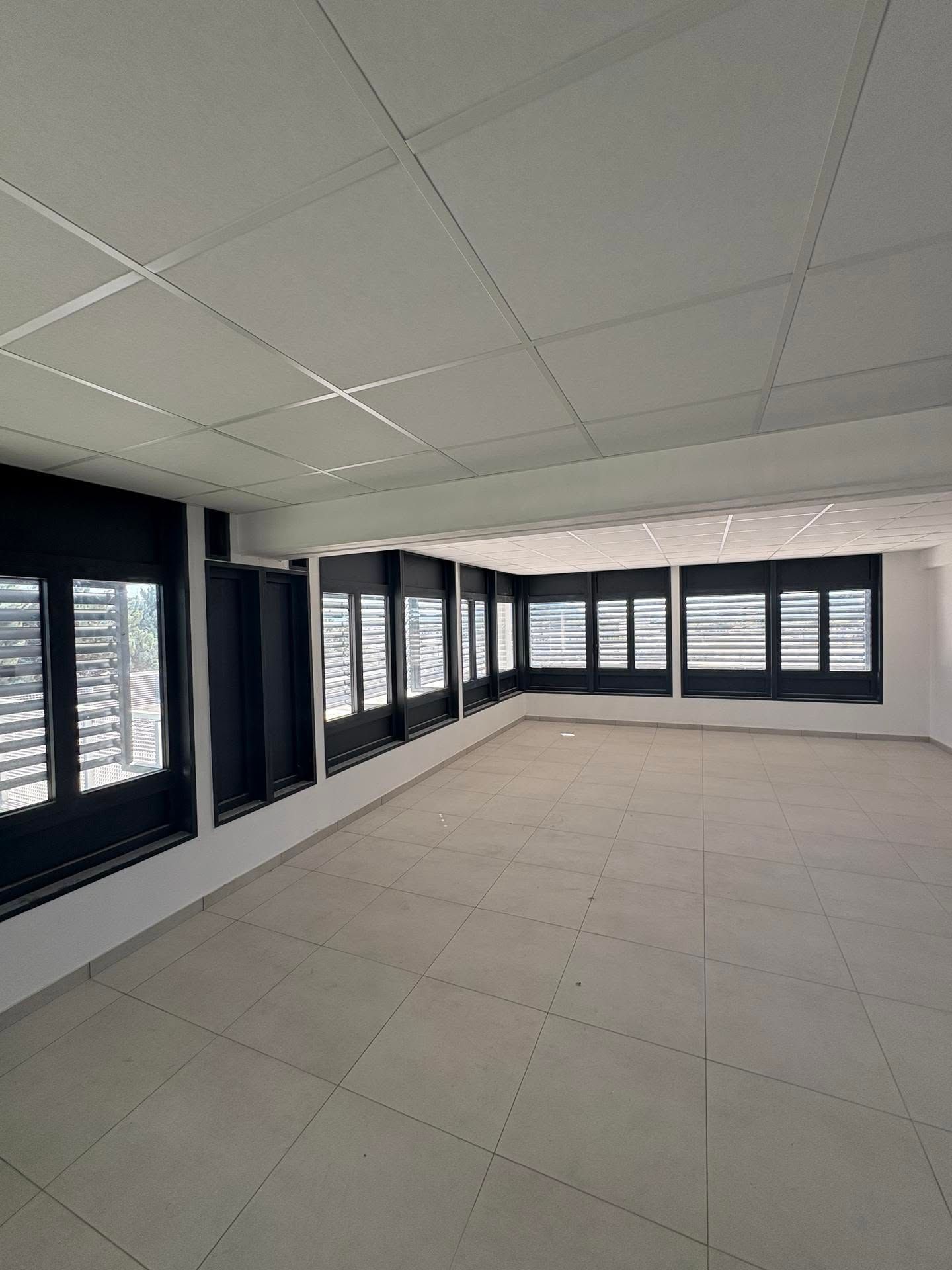 Espace de bureau moderne vide avec de grandes fenêtres, des dalles de plafond blanches et un sol carrelé clair.