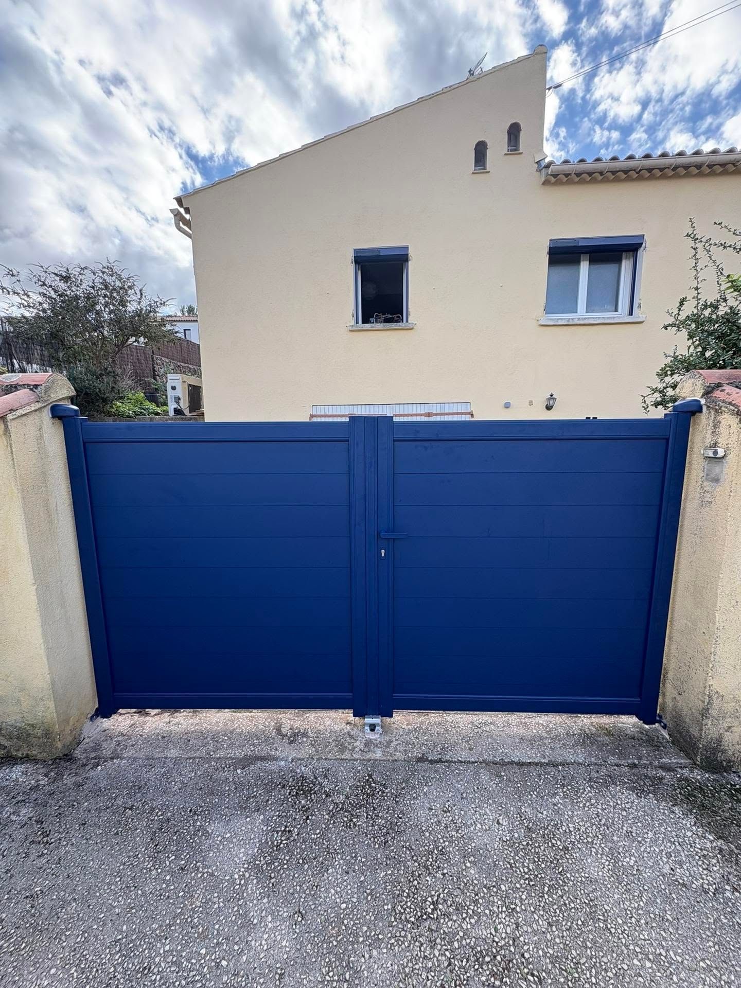 Un portail en métal bleu devant une maison beige, sous un ciel nuageux.