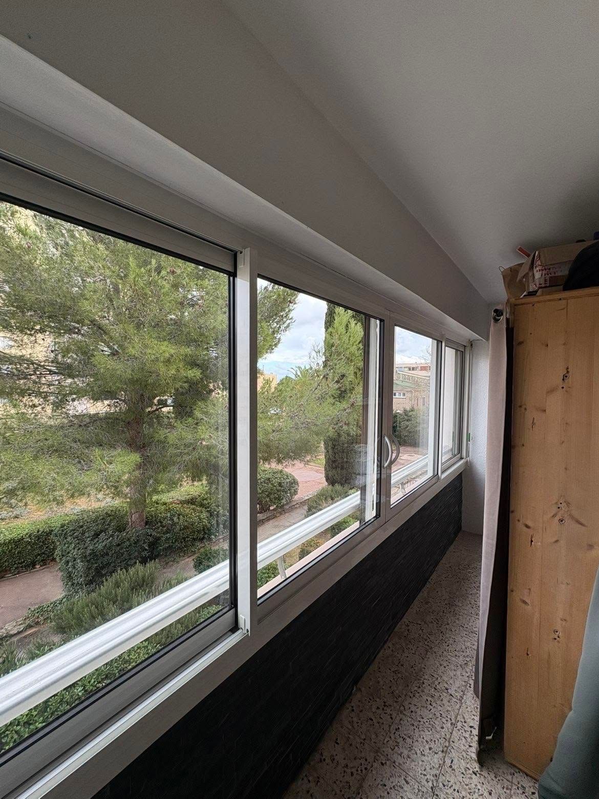 Couloir intérieur étroit avec de grandes fenêtres donnant sur des arbres et une haie, à côté d'une armoire en bois.