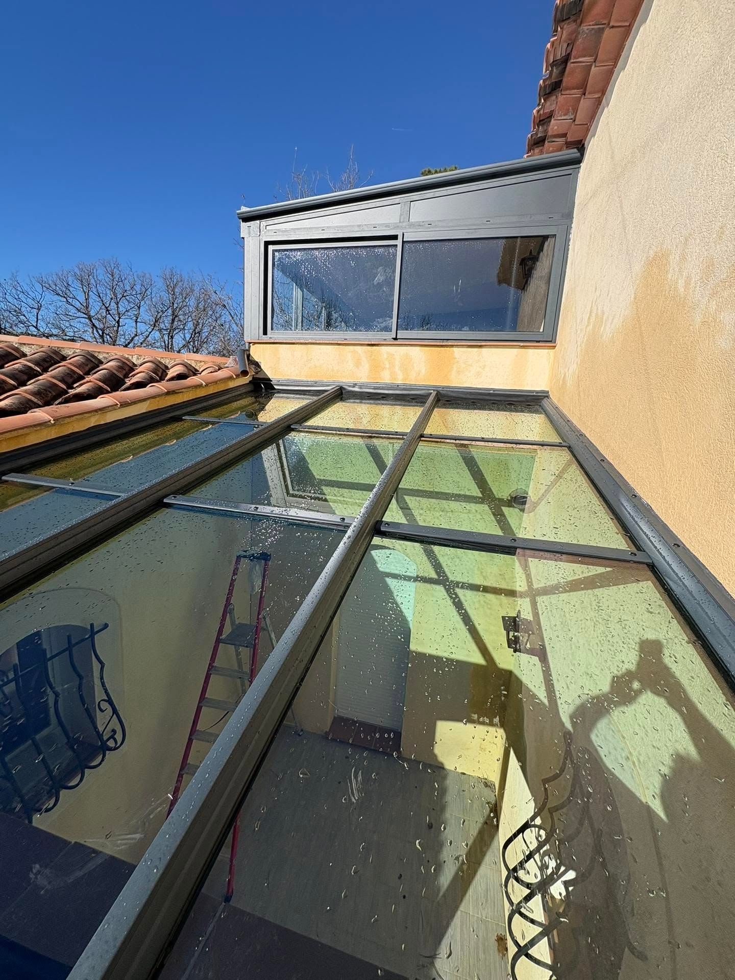 Terrasse ou véranda à toit de verre, vue vers le haut entre des murs jaunes et gris, avec un ciel bleu au-dessus.