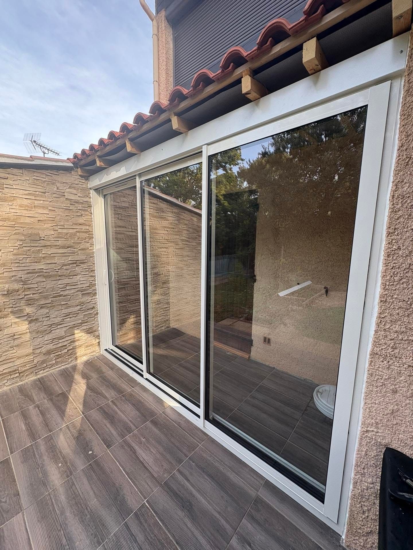 Des portes coulissantes en verre donnent sur une terrasse en bois sous un toit de tuiles, reflétant les arbres et le ciel.
