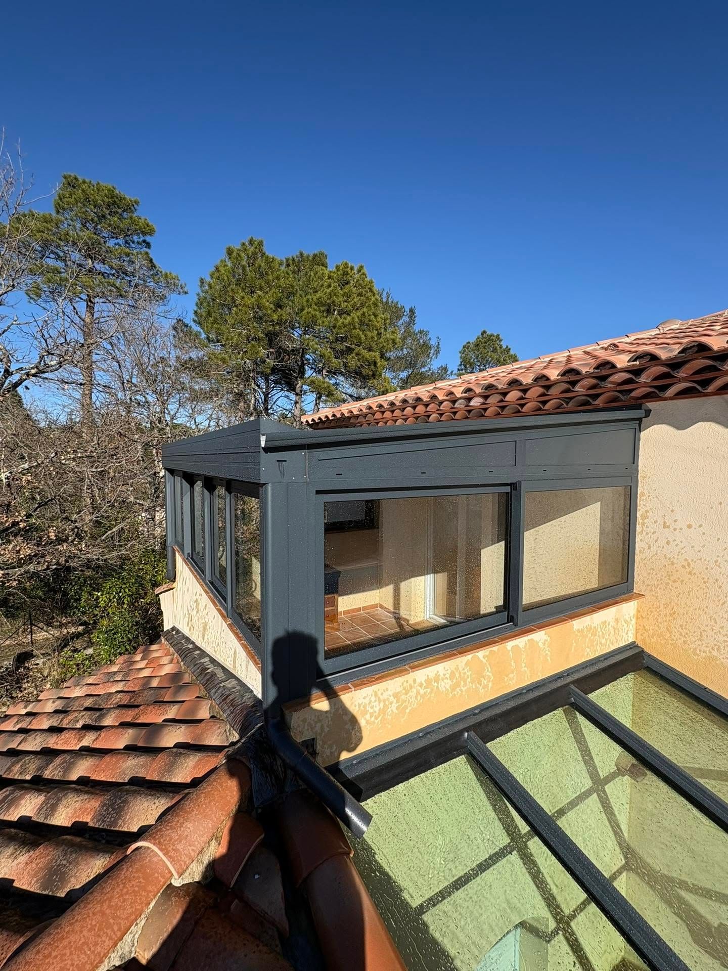 Véranda vitrée sur le toit, avec toit de tuiles rouges et arbres sous un ciel bleu azur.