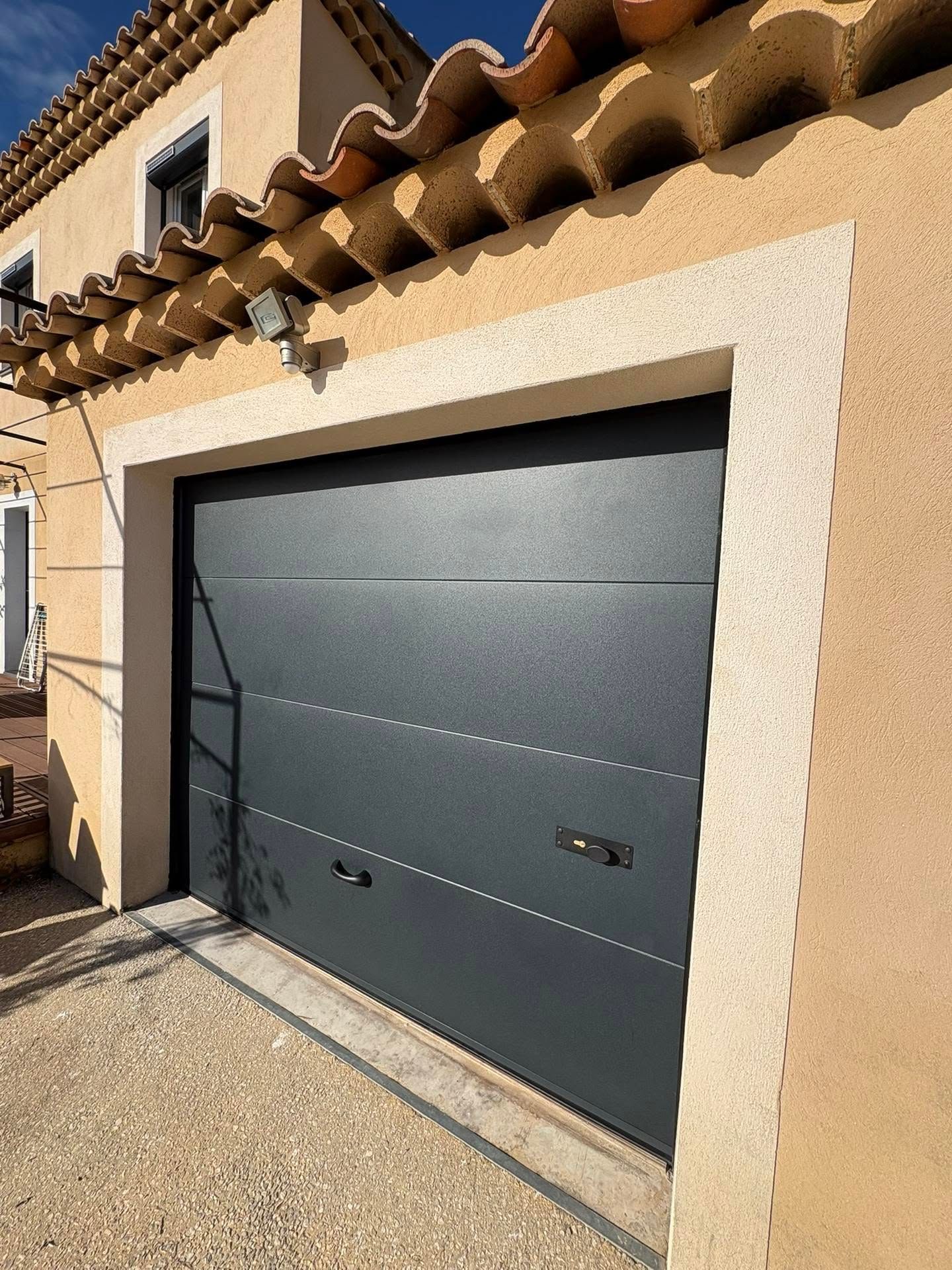 Porte de garage grise moderne sur une maison en stuc beige avec un auvent carrelé, baignée de soleil.