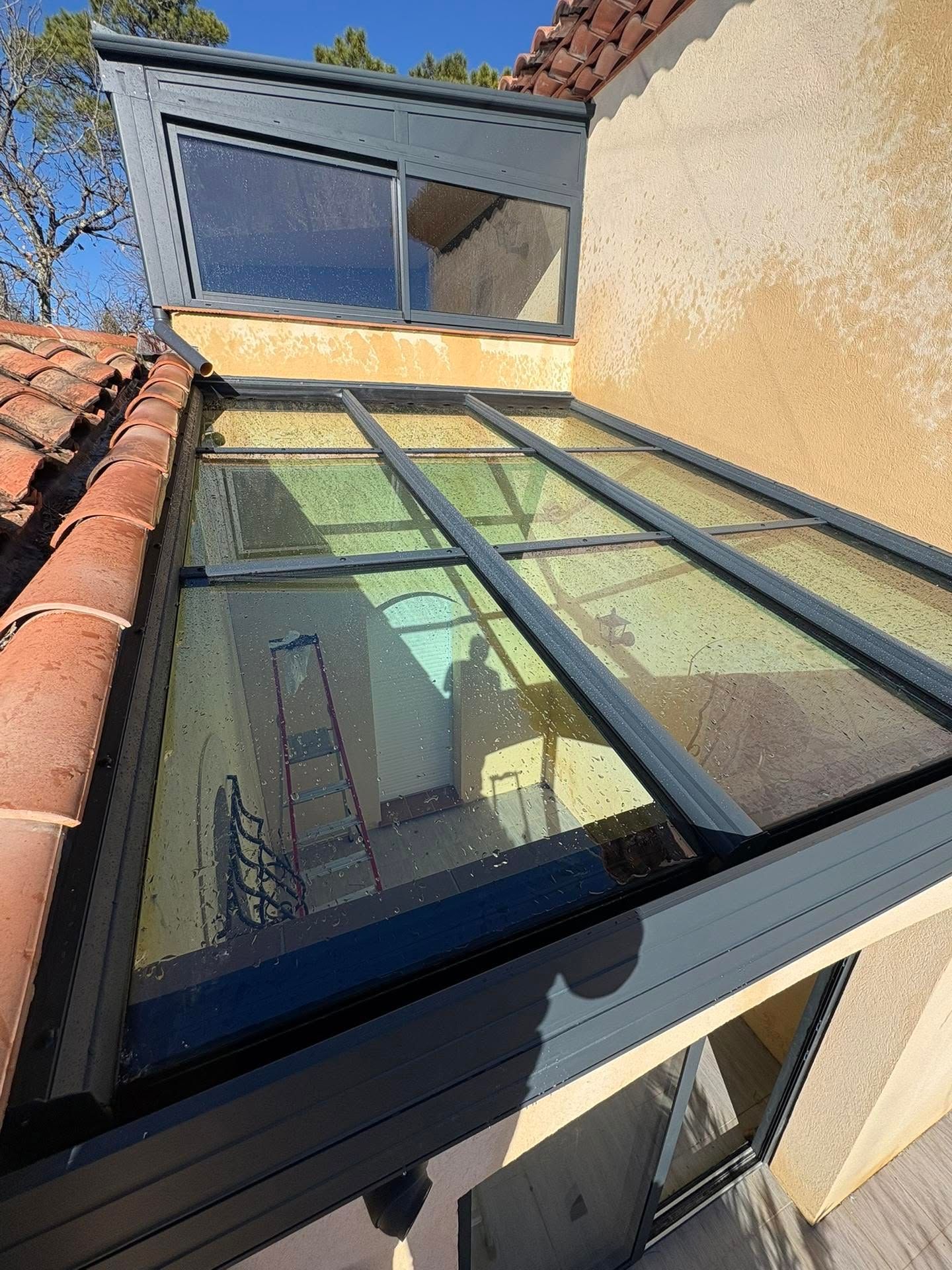 Verrière en verre sur le toit d'un bâtiment en stuc, reflétant le ciel bleu à côté des tuiles de terre cuite rouges.