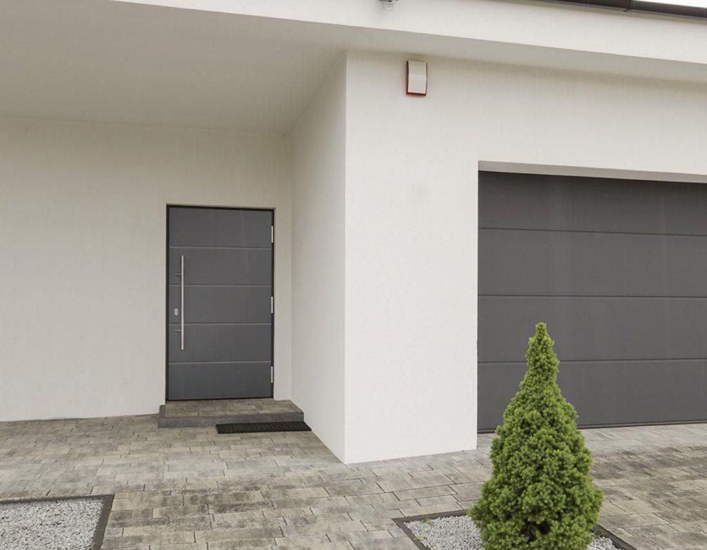 Entrée de maison moderne avec porte grise, porte de garage et petit arbuste à feuillage persistant sur une terrasse en pierre.